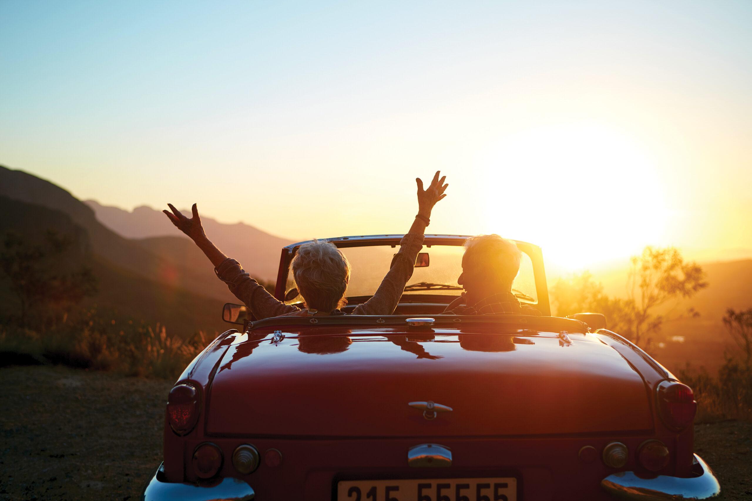 A happy couple drives into the sunset in an open convertible. The person to the left has their arms outstretched.