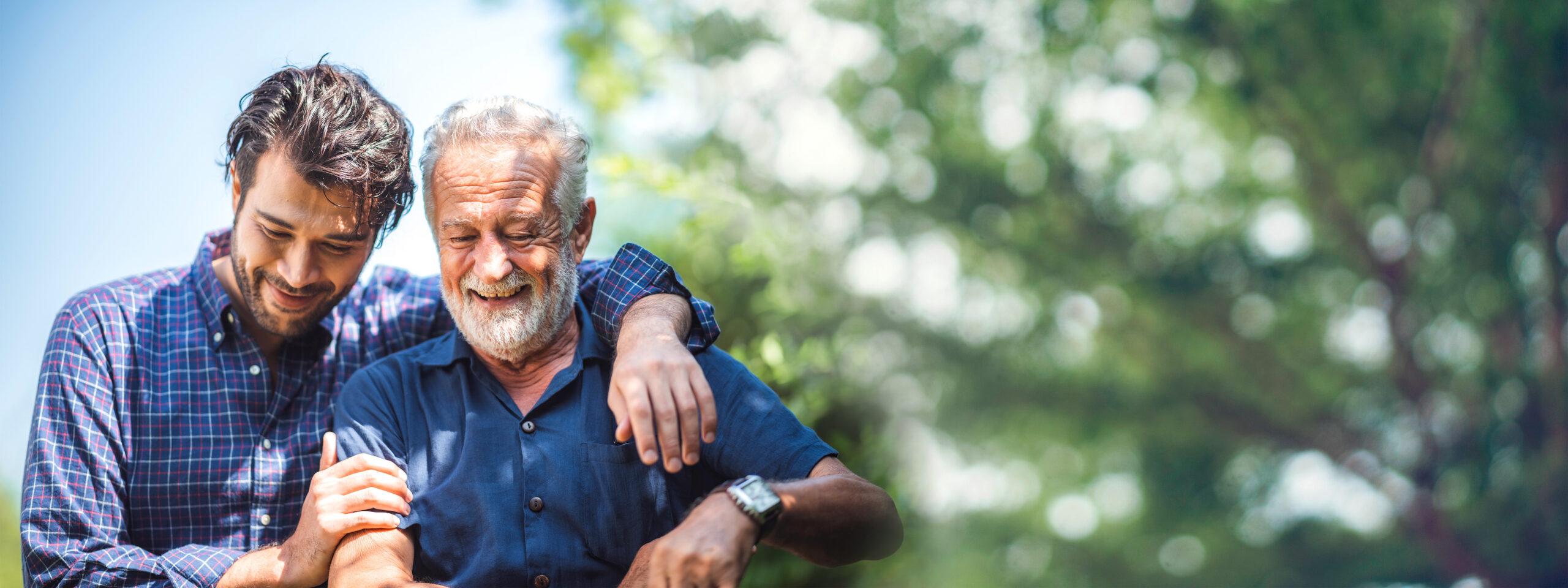 An adult son smiles with his arm around his father outdoors.